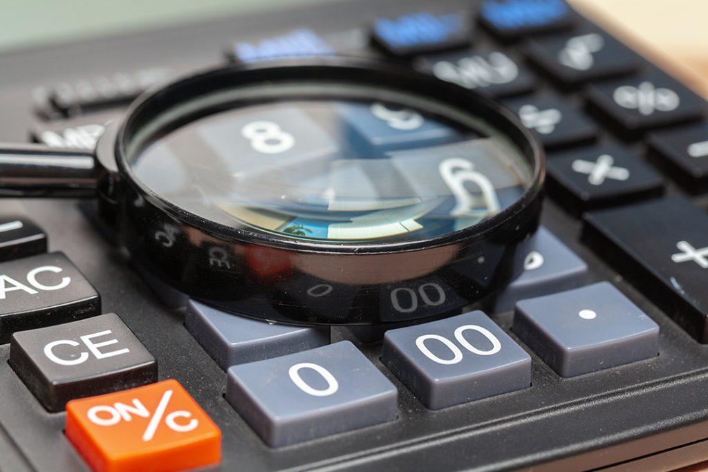 Lupa sobre uma calculadora preta com as teclas azuis, cinzas e vermelha para analisar um fechamento contábil.